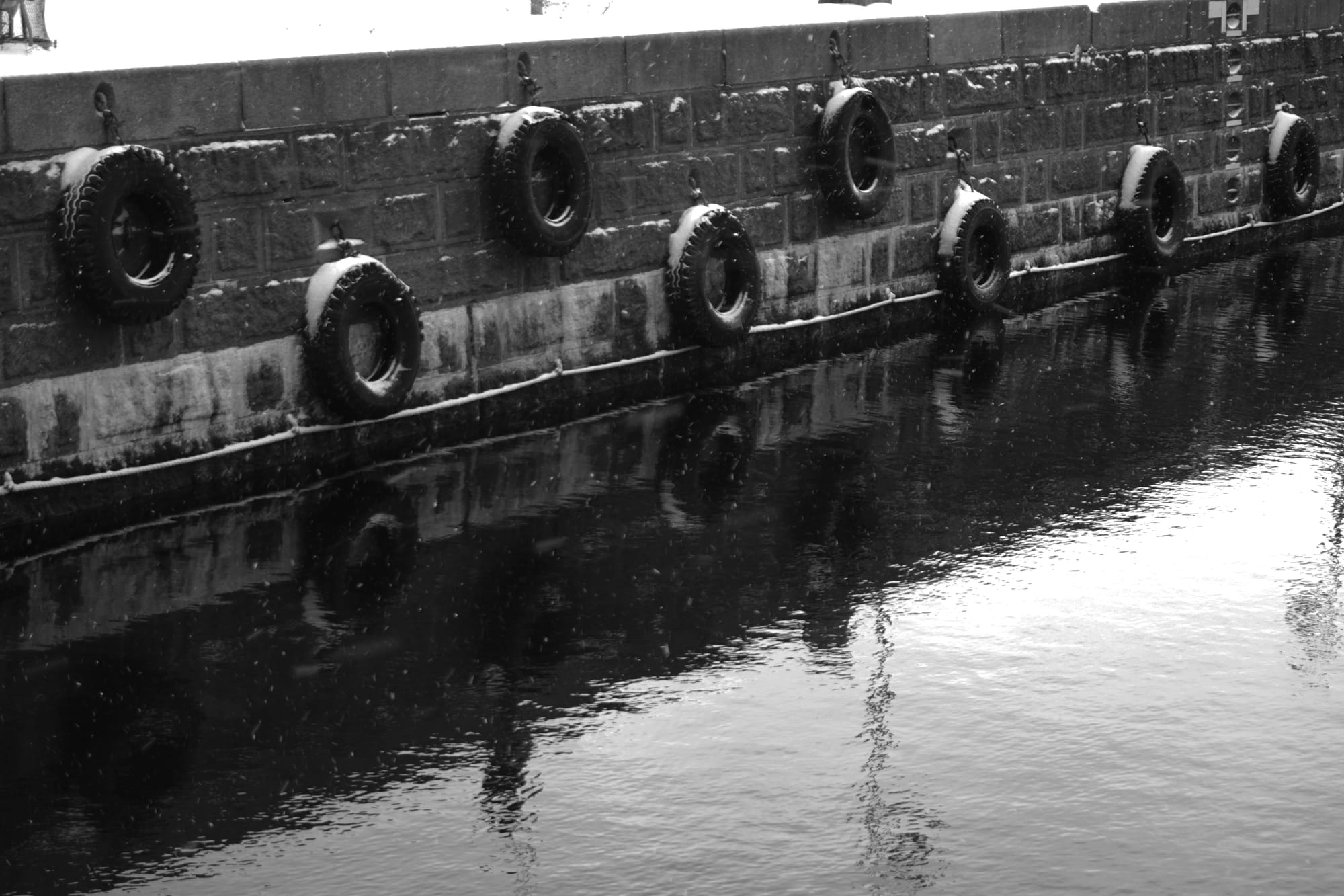 Black and white photo of tyres on the side of a lock. Snow sits atop the tyres.