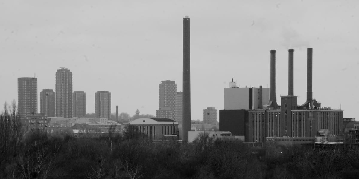 A view of a skyline, with tall apartment buildings, and a factory with chimneys. Winter trees fill the bottom third of the image in the foreground.